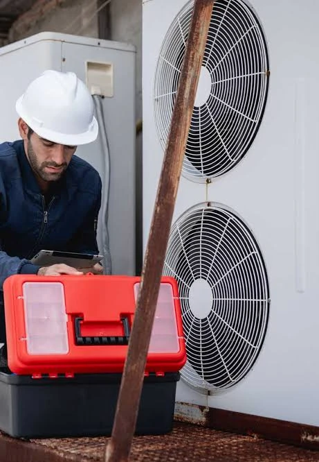 Technician in a hard hat inspecting a large outdoor HVAC unit with a tablet and toolbox